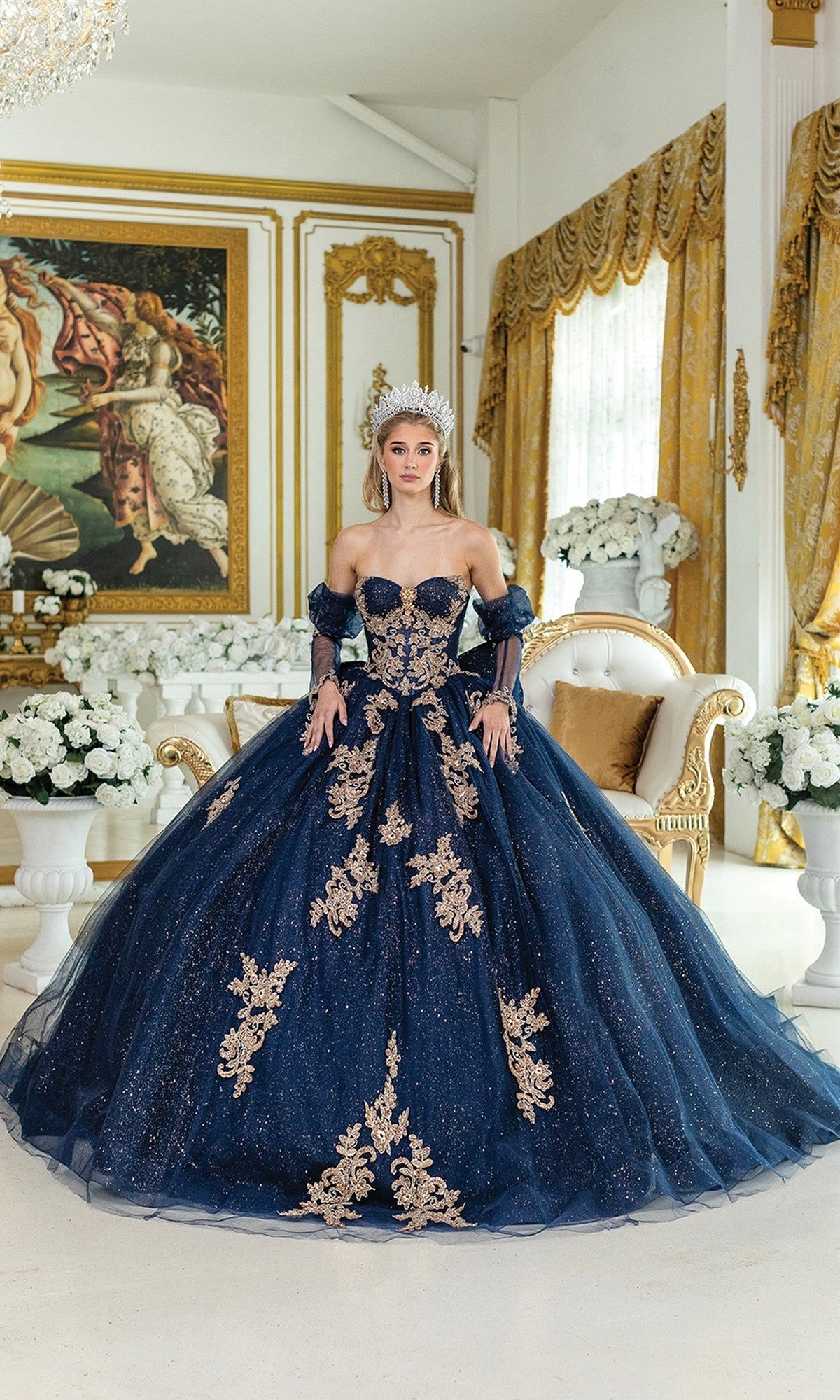 A woman wearing the Quinceanera Dress 5051 By Dancing Queen—a navy blue and gold embellished glitter tulle ball gown with a tiara—stands in an ornate room with gold accents, white flowers, and a grand classical painting.